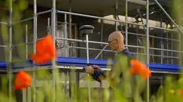 Ein Vermessungsingenieur arbeitet mit einem GPS-Messgerät vor einem eingerüsteten Gebäude, im Vordergrund blühen orangefarbene Mohnblumen