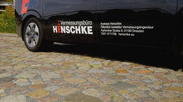 Die Seitenansicht eines schwarzen Fahrzeugs mit der Aufschrift „Vermessungsbüro Henschke“, geparkt auf Kopfsteinpflaster, zeigt die Kontaktdaten des öffentlich bestellten Vermessungsingenieurs Andreas Henschke aus Dresden