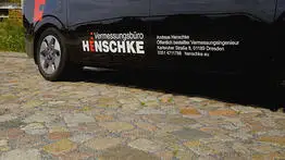 Die Seitenansicht eines schwarzen Fahrzeugs mit der Aufschrift „Vermessungsbüro Henschke“, geparkt auf Kopfsteinpflaster, zeigt die Kontaktdaten des öffentlich bestellten Vermessungsingenieurs Andreas Henschke aus Dresden