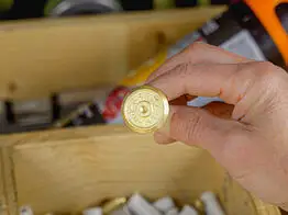 Eine Hand hält eine messingfarbene Grenzmarke mit der Aufschrift „Grenzpunkt“ über einem Fach im Werkzeugkasten, in dem weitere Kunststoffhülsen und Arbeitsmaterialien liegen