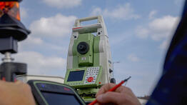 Eine grüne Leica-Totalstation steht auf einem Stativ unter blauem Himmel, im Vordergrund hält eine Hand ein Datenerfassungsgerät mit rotem Stift und Reflektor