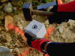 Eine Person mit roten Handschuhen schlägt mit einem Hammer einen weißen Grenzpunkt-Marker in Dresden mit der Aufschrift „Grenzpunkt“ in den Boden ein, im Hintergrund ist orangefarbene Markierfarbe sichtbar
