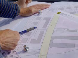 Vermessungsingenieuer aus Dresden zeigt mit einem Kugelschreiber auf einen Plan mit Gebäuden und Grundstücksgrenzen