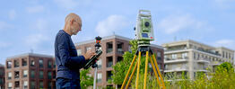 Vermessungsingenieur Andreas Henschke misst mit einer Totalstation und einem Reflektorstab auf einem Gelände vor modernen Wohnhäusern