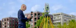 Vermessungsingenieur Andreas Henschke misst mit einer Totalstation und einem Reflektorstab auf einem Gelände vor modernen Wohnhäusern