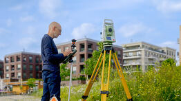 Vermessungsingenieur Andreas Henschke misst mit einer Totalstation und einem Reflektorstab auf einem Gelände vor modernen Wohnhäusern