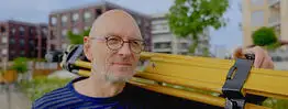 Vermessungsingenieur und Gutachter Andreas Henschke aus Dresden trägt ein gelbes Stativ auf der Schulter, im Hintergrund sind moderne Wohngebäude und Grünflächen zu sehen
