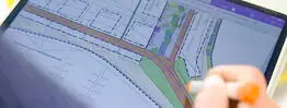 Eine Hand bearbeitet mit einem digitalen Stift einen farbigen Stadtplan auf einem Tablet, sichtbar sind geplante Straßen und Grünflächen.