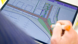 Eine Hand bearbeitet mit einem digitalen Stift einen farbigen Stadtplan auf einem Tablet, sichtbar sind geplante Straßen und Grünflächen.