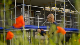 Vermessungsingenieur Andreas Henschke arbeitet mit einem GPS-Messgerät vor einem eingerüsteten Gebäude, im Vordergrund blühen orangefarbene Mohnblumen