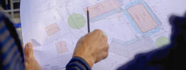 Eine Hand zeigt mit einem Bleistift auf einen detaillierten Lageplan mit Grundstücksgrenzen und Gebäudeflächen