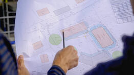 Eine Hand zeigt mit einem Bleistift auf einen detaillierten Lageplan mit Grundstücksgrenzen und Gebäudeflächen