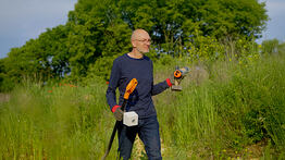 Vermessungsingenieur Andreas Henschke mit Werkzeugen in den Händen steht in einem grünen, bewachsenen Gelände und bereitet eine Messung vor
