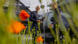 Vermessungsingenieur Andreas Henschke begrüßt einen privaten Bauherren vor einem Vermessungsfahrzeug auf einer Baustelle mit Gerüst, im Vordergrund blühen orangefarbene Mohnblumen