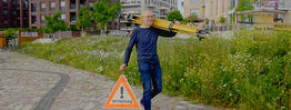 Vermessungsingenieur Andreas Henschke trägt ein gelbes Stativ über der Schulter und ein Warndreieck mit der Aufschrift „Vermessung“ in der Hand, während er über einen gepflasterten Weg in einem modernen Stadtviertel geht