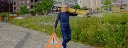 Vermessungsingenieur Andreas Henschke trägt ein gelbes Stativ über der Schulter und ein Warndreieck mit der Aufschrift „Vermessung“ in der Hand, während er über einen gepflasterten Weg in einem modernen Stadtviertel geht