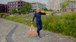 Vermessungsingenieur Andreas Henschke trägt ein gelbes Stativ über der Schulter und ein Warndreieck mit der Aufschrift „Vermessung“ in der Hand, während er über einen gepflasterten Weg in einem modernen Stadtviertel geht