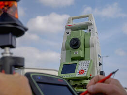 Eine grüne Leica-Totalstation steht auf einem Stativ unter blauem Himmel, im Vordergrund hält eine Hand ein Datenerfassungsgerät mit rotem Stift und Reflektor