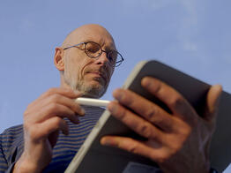 Ein Vermessungsingenieur blickt konzentriert auf ein Tablet und hält einen Eingabestift in der Hand. Der Himmel im Hintergrund ist blau