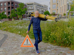 Ein Vermessungsingenieur trägt ein gelbes Stativ über der Schulter und ein Warndreieck mit der Aufschrift „Vermessung“ in der Hand, während er über einen gepflasterten Weg in einem modernen Stadtviertel geht