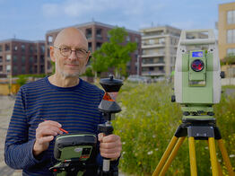 Vermessungsingenieur Andreas Henschke steht mit Prisma und Steuergerät auf einem Pflasterweg, neben ihm eine Totalstation auf Stativ; im Hintergrund moderne Wohngebäude und eine Blumenwiese