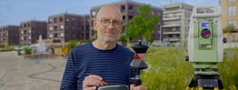 Vermessungsingenieur Andreas Henschke steht mit Prisma und Steuergerät auf einem Pflasterweg, neben ihm eine Totalstation auf Stativ; im Hintergrund moderne Wohngebäude und eine Blumenwiese
