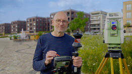 Vermessungsingenieur Andreas Henschke steht mit Prisma und Steuergerät auf einem Pflasterweg, neben ihm eine Totalstation auf Stativ; im Hintergrund moderne Wohngebäude und eine Blumenwiese